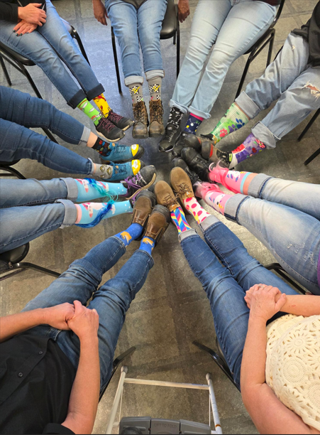 A group of people sitting in a circle with their feet pointed inward, showcasing bright, colorful, and mismatched socks in honor of World Down Syndrome Day.