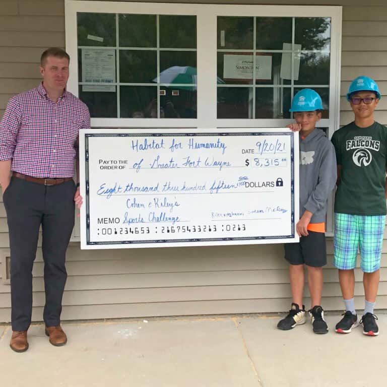 Checkpresentation of large donation check for Habitat for Humanity by Kelco Industries employees and local youth in safety helmets in front of a building, promoting community support and corporate responsibility.