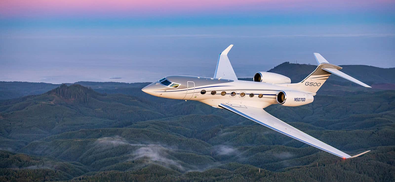 Sleek private jet flying over lush green mountains at sunrise or sunset.