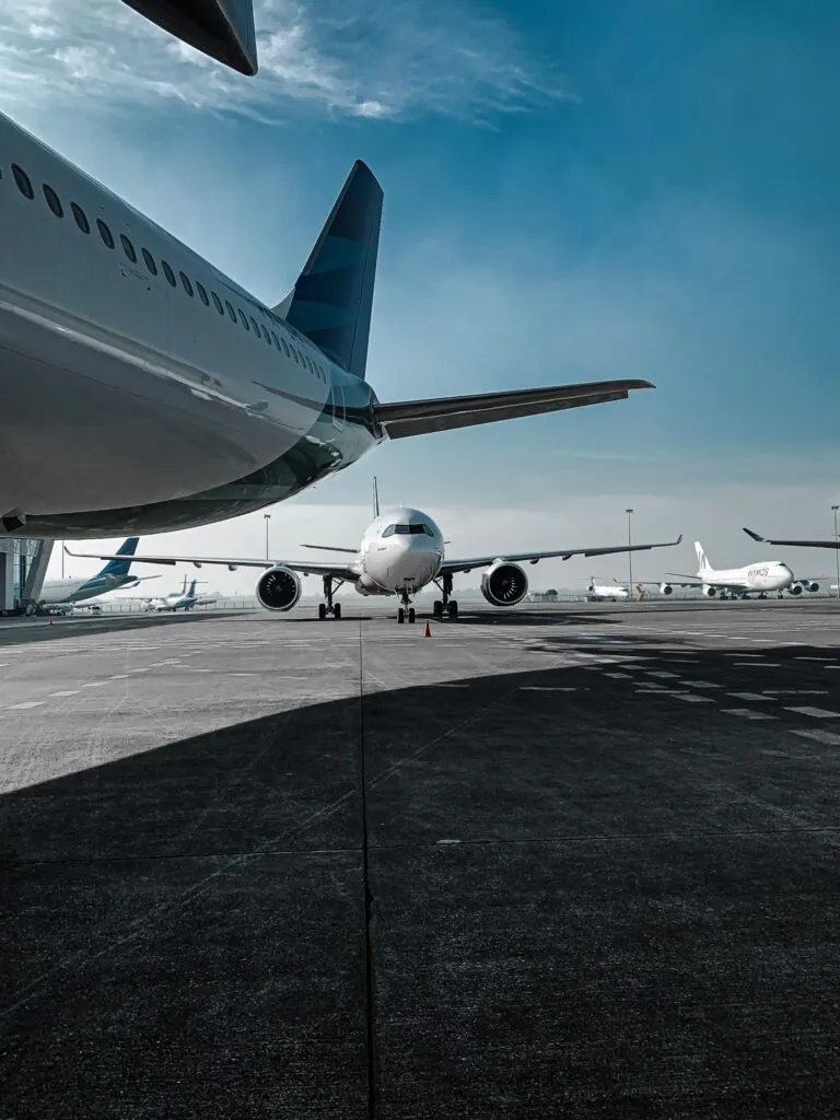 A row of commercial airplanes parked on the tarmac at an airport. The perspective shows the tail and fuselage of one aircraft prominently in the foreground, with another plane directly ahead and more aircraft visible in the distance, illustrating the bustling activity and scale of modern air travel.