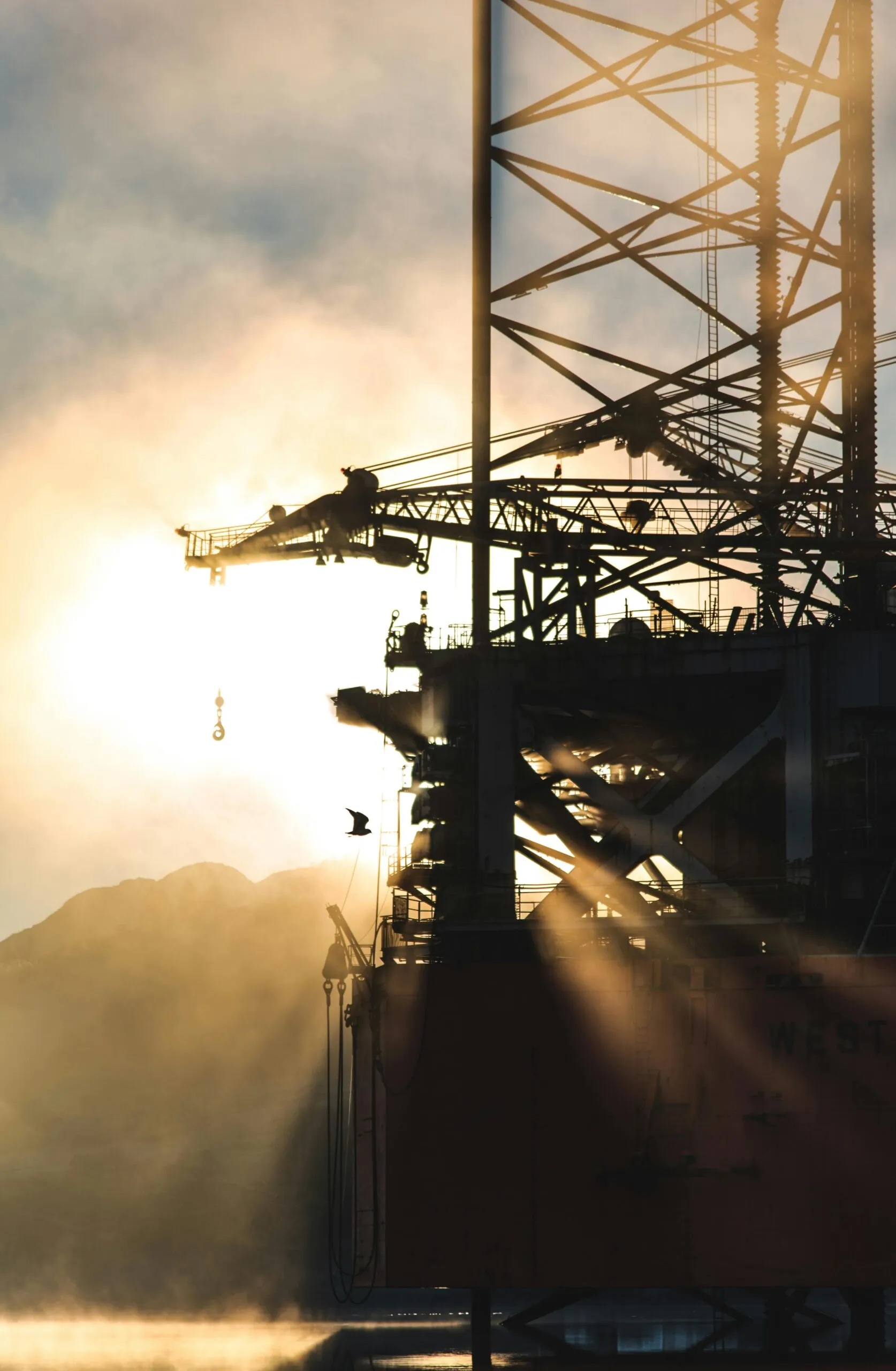 Silhouetted offshore oil rig with intricate structural details against the backdrop of a misty sunrise. The sun's rays and mist create a dramatic effect, highlighting the rig's complex machinery and a bird flying nearby, capturing the serene yet industrious nature of offshore drilling operations.