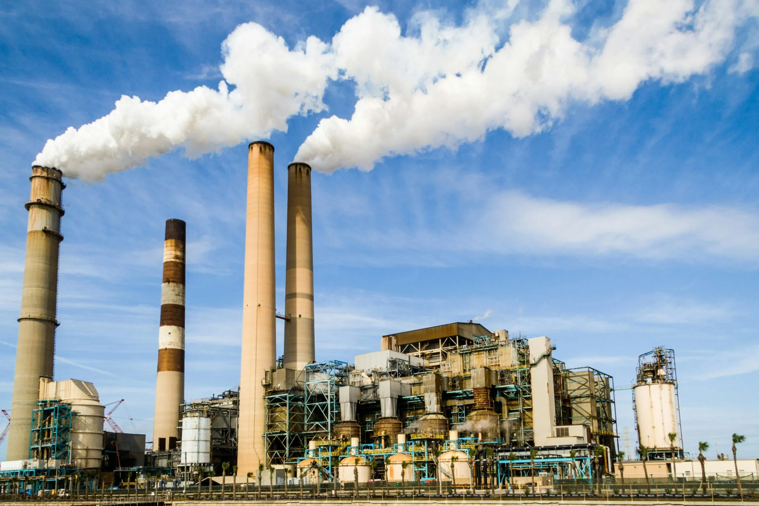 An industrial power plant with multiple tall smoke stacks emitting white steam against a blue sky. The facility is a complex structure of pipes, tanks, and machinery, highlighting the scale and infrastructure involved in industrial power generation.