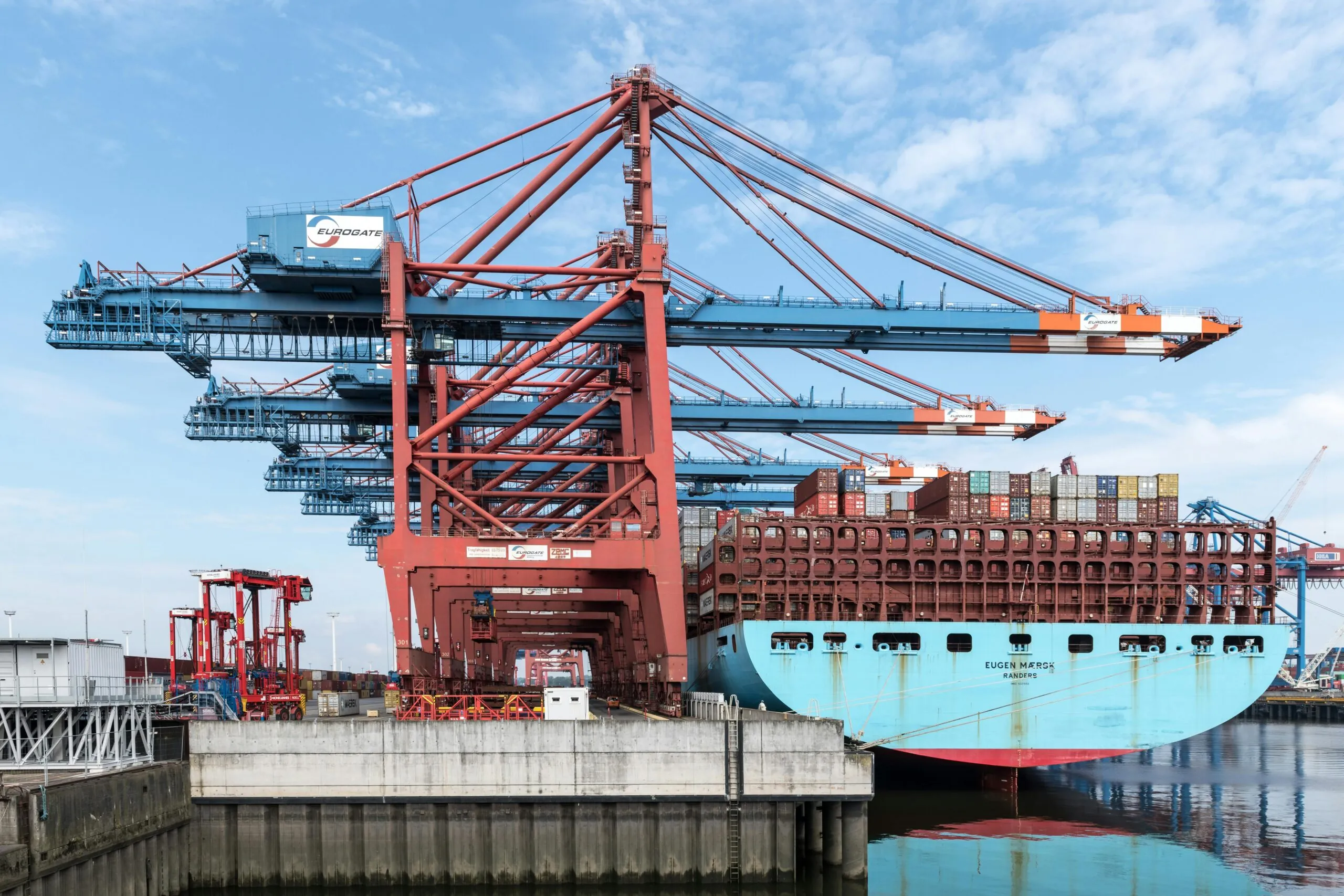 A large container ship docked at a port, with towering red and blue loading cranes in the background. The cranes, belonging to Eurogate, are used for loading and unloading shipping containers, highlighting the scale and efficiency of modern maritime logistics.