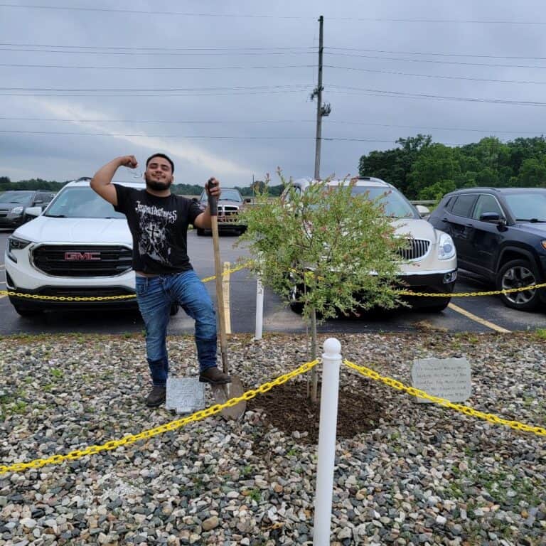 Young man planting a tree in a parking lot with cars behind, symbolizing environmental initiatives and urban forestry.